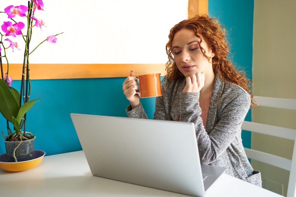 Beautiful redhead woman teleworking on laptop at home and drinking coffee.